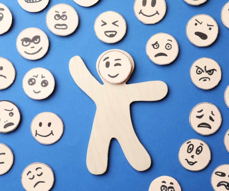 Obrazek wyróżniający do artykułu poświęconego emocjom. Emocje - rodzaje, funkcje, lista emocji. Ludzik na niebieskim tle, dookoła niego buźki z różnymi emocjami.