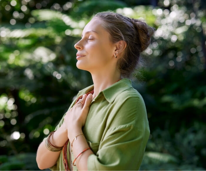 Kobieta trzymająca dłonie na piersi i wsłuchująca się w swoje wnętrze. Na łonie natury. Przyswaja najpiękniejsze cytaty o emocjach.